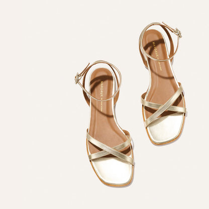 The Flat Sandal - Champagne by Margaux features gold strappy flats with ankle straps and a foam-cushioned insole, shown on a light beige background for timeless style and comfort.