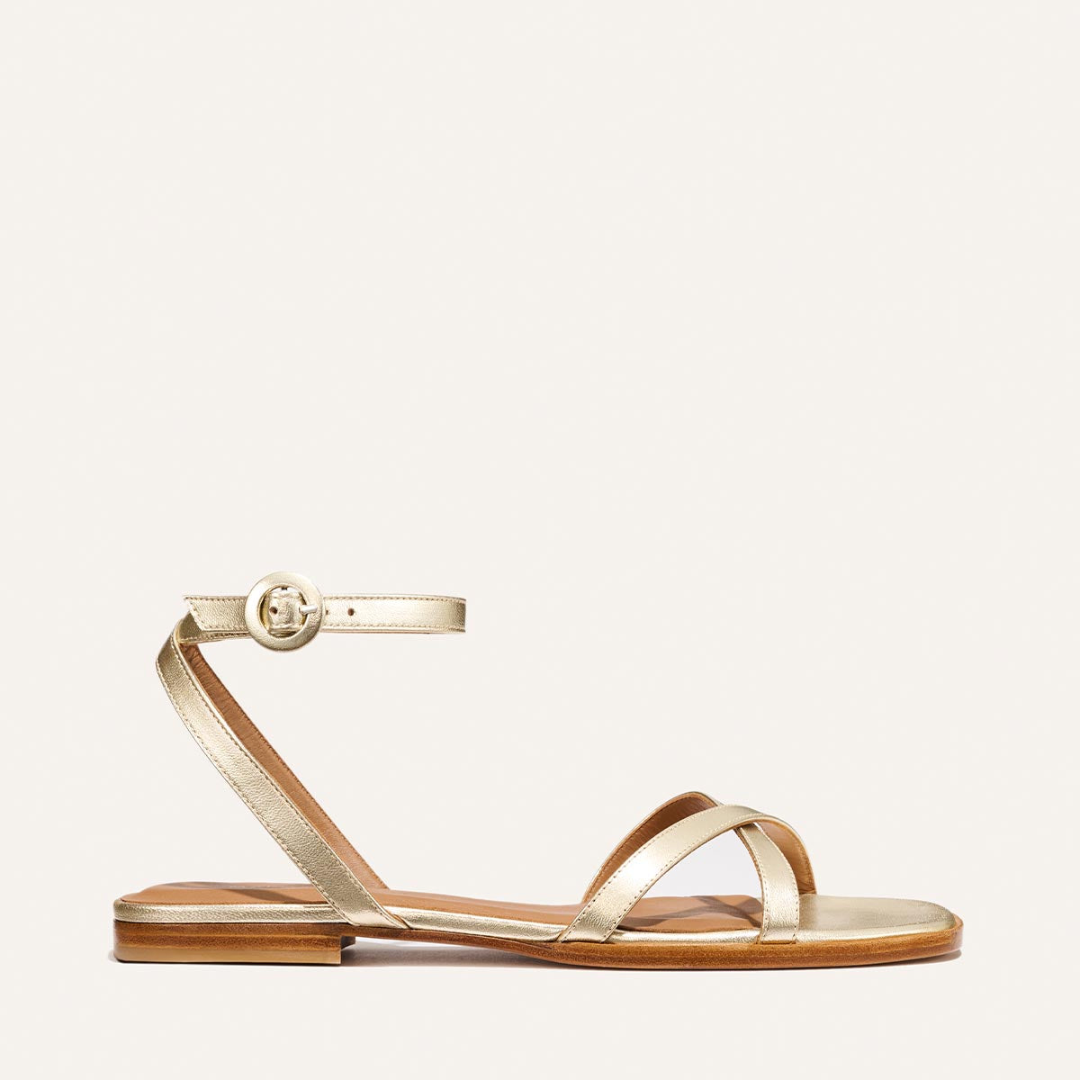 The Flat Sandal - Champagne by Margaux: a gold barely-there style with thin straps, an ankle strap with round buckle, flat brown sole, and foam-cushioned insole for comfort. Shown on a plain white background.