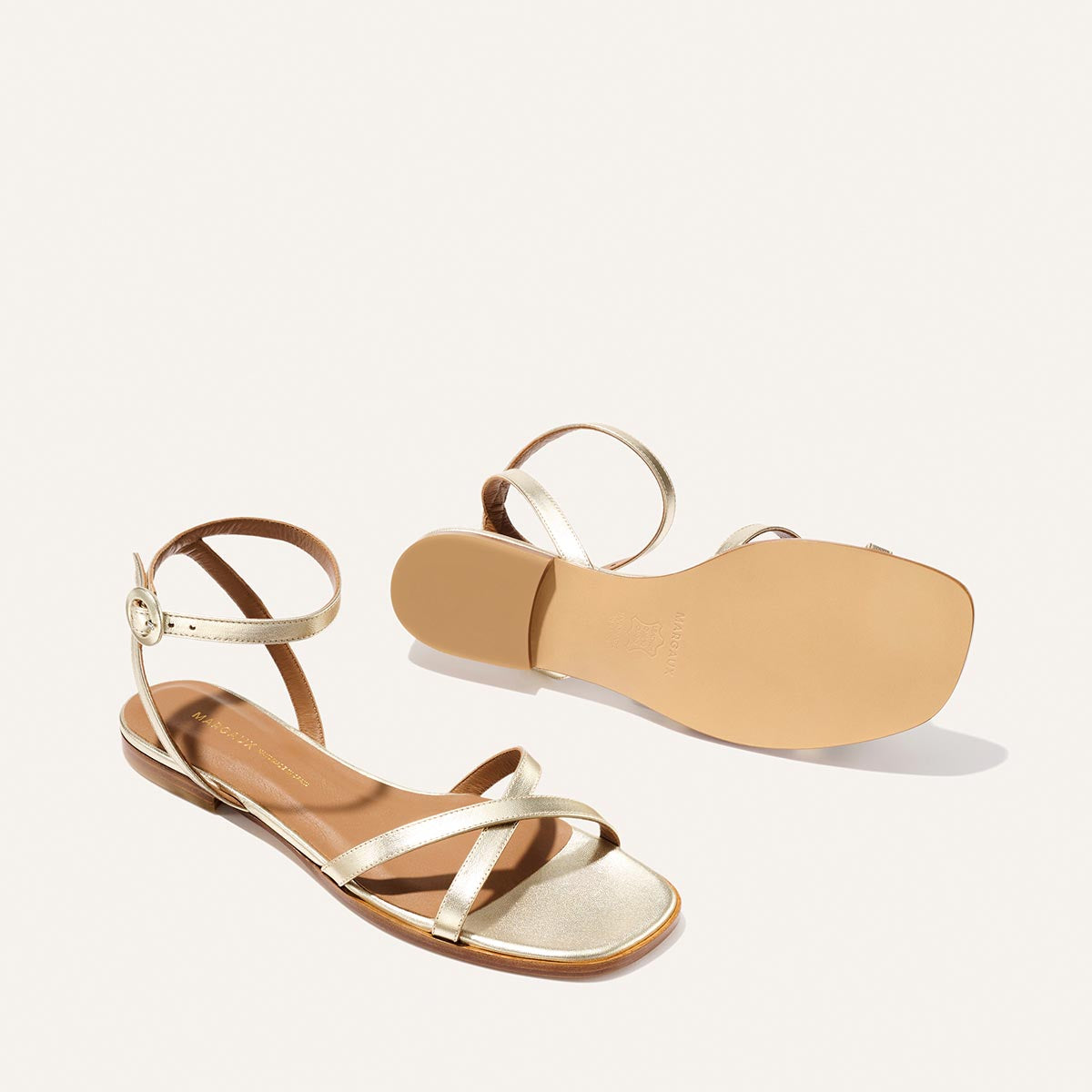 The Flat Sandal - Champagne by Margaux is shown on a white background, featuring gold strappy design, ankle straps with buckle closures, and a foam-cushioned insole for a barely-there feel. One sandal displays the sole.