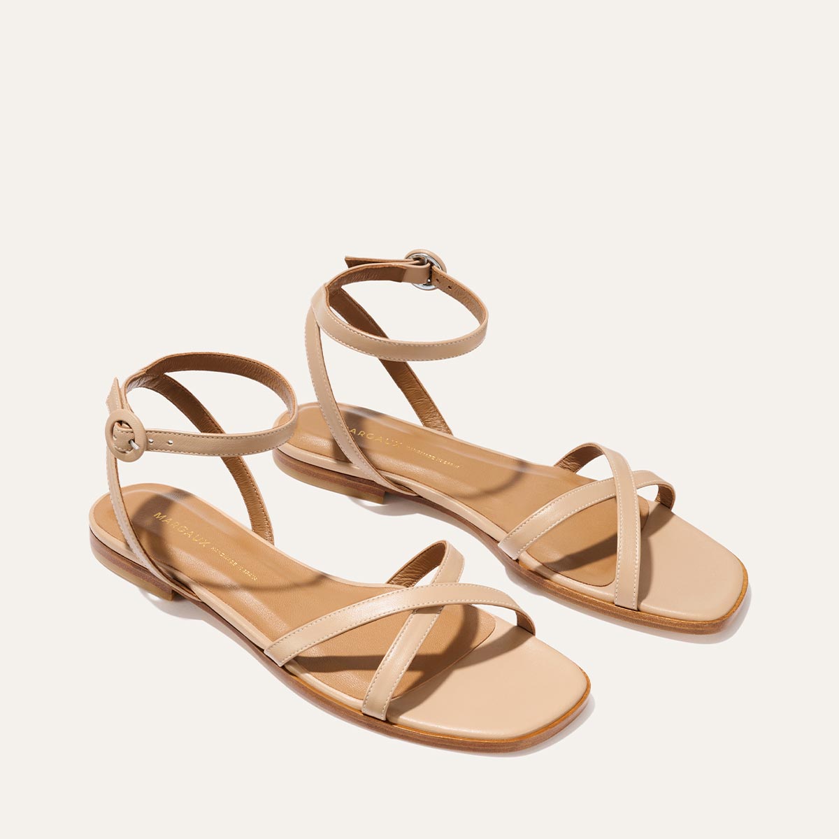 The Margaux Flat Sandal - Dune Nappa features thin crisscross toe straps, adjustable ankle straps, and a foam-cushioned insole for all-day comfort, shown in beige on a light background.