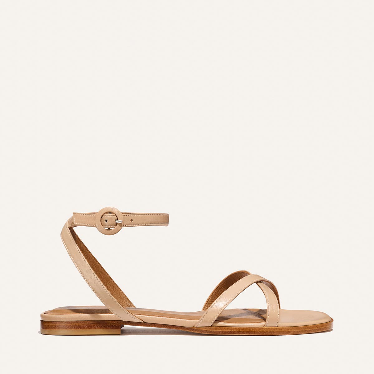 The Flat Sandal - Dune Nappa by Margaux is a tan barely-there sandal featuring a thin ankle strap with buckle, two crossing toe straps, foam-cushioned insole, and low wooden sole, shown on a plain light background.