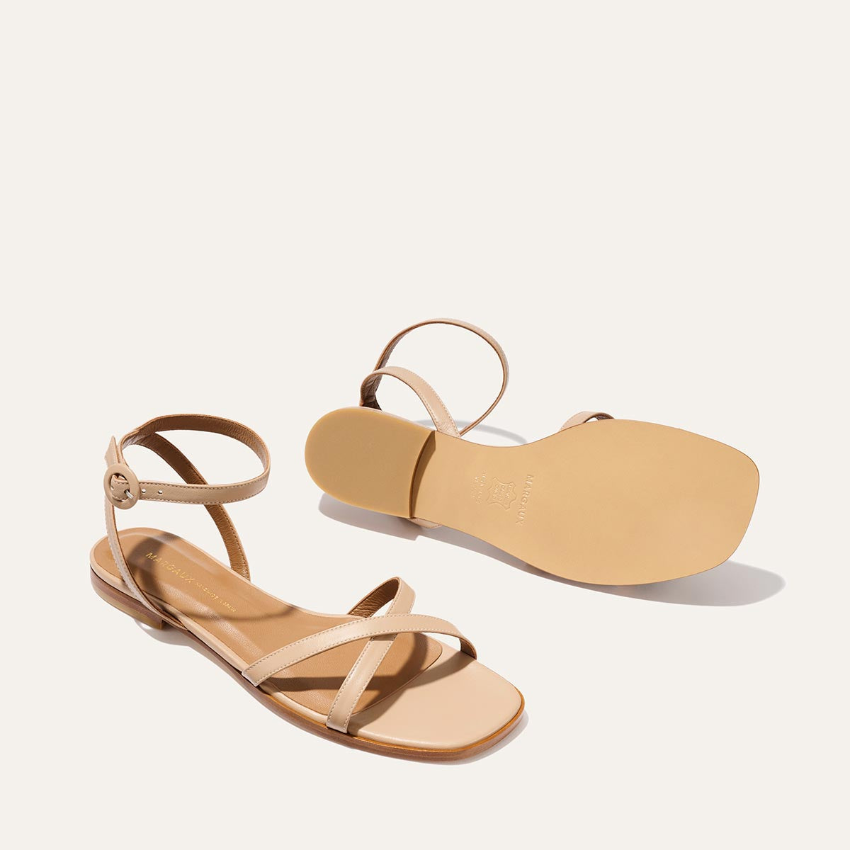 The Margaux Flat Sandal - Dune Nappa: a beige pair with ankle straps, flat soles, and foam-cushioned insoles. One sandal stands upright; the other lays on its side to showcase the sole, all against a light background.