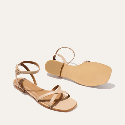 The Margaux Flat Sandal - Dune Nappa: a beige pair with ankle straps, flat soles, and foam-cushioned insoles. One sandal stands upright; the other lays on its side to showcase the sole, all against a light background.