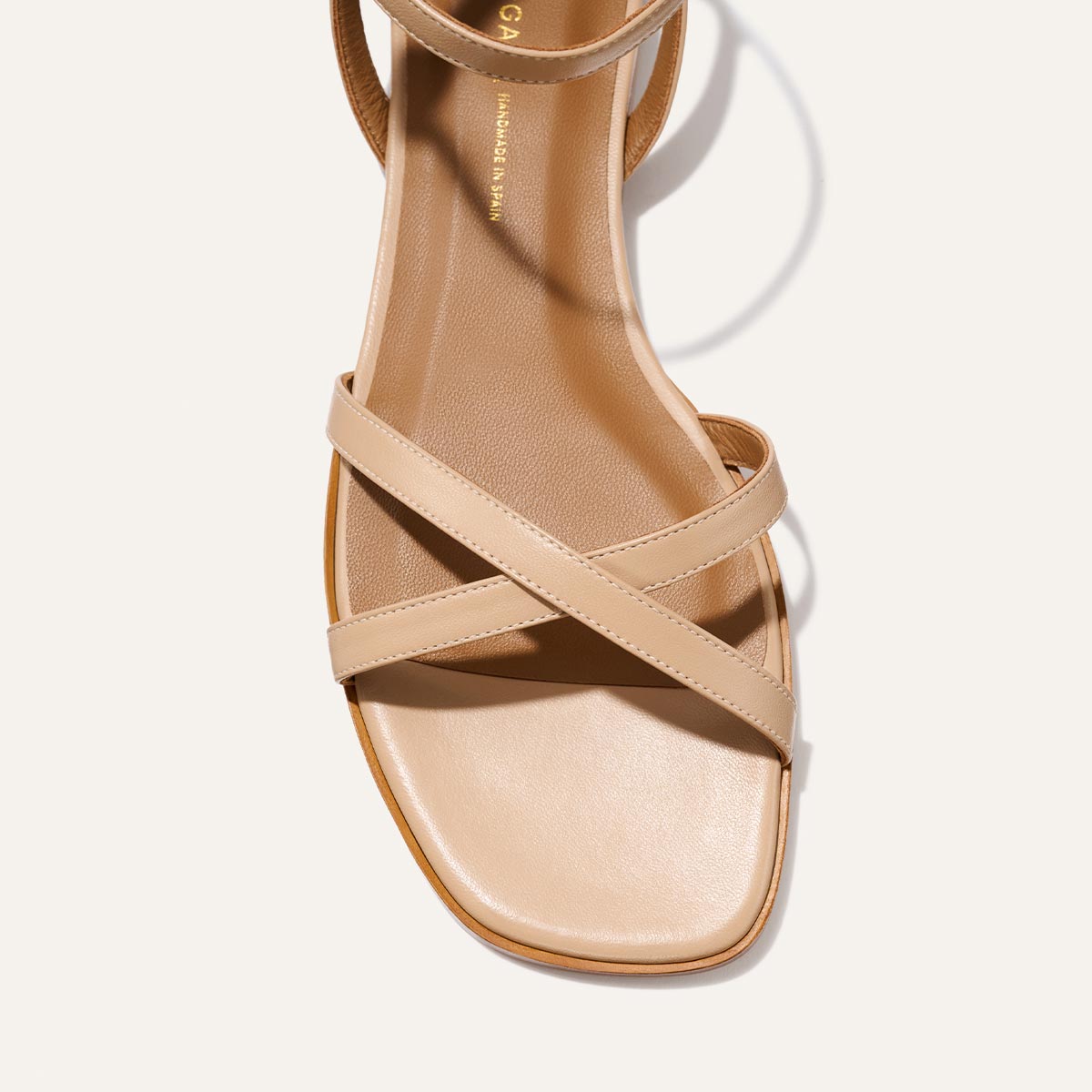 The Flat Sandal - Dune Nappa by Margaux features a beige, barely-there design with a square toe, two slim crisscross straps, and a foam-cushioned insole for comfort. Photographed from above on a light background.