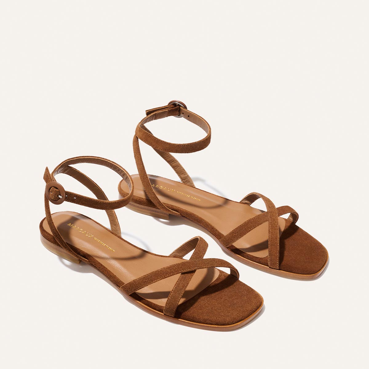 The Flat Sandal - Saddle Suede by Margaux features brown suede, cushioned insoles, open toes, crisscross front straps, and ankle straps, shown on a white background.