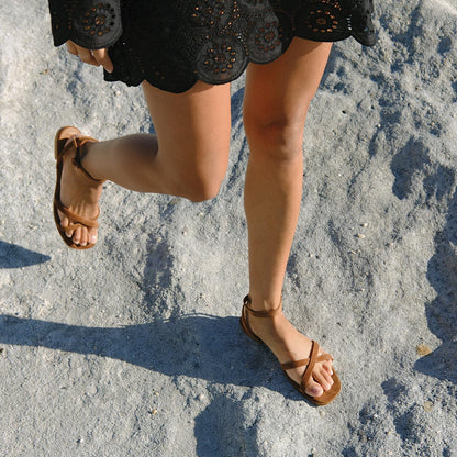 A person in a black eyelet dress and Margaux's The Flat Sandal in Saddle Suede walks across sunlit rocks, showing only their legs and feet.