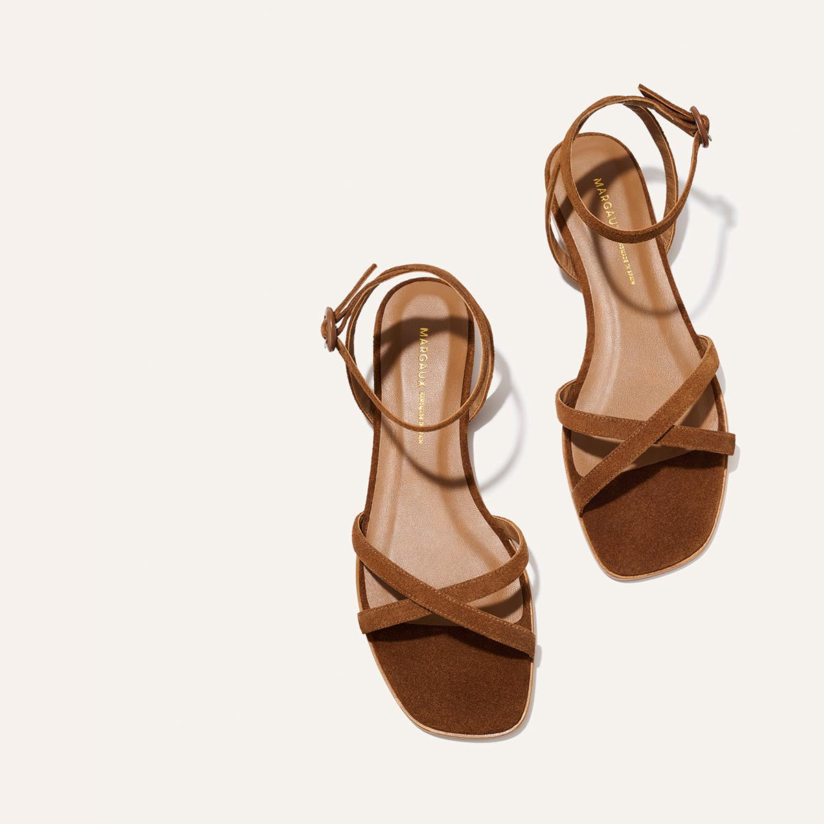 The Margaux Flat Sandal in Saddle Suede features ankle straps, crisscross toe straps, and a foam-cushioned insole for comfort and timeless style, elegantly displayed on a light background.