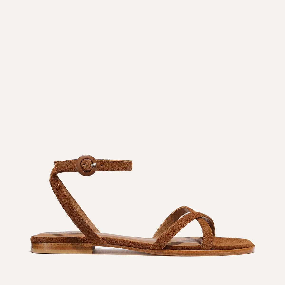 The Flat Sandal - Saddle Suede by Margaux is a brown suede barely-there sandal with slim crossing straps, a thin ankle strap and buckle, flat sole, and foam-cushioned insole, shown against a plain light background.