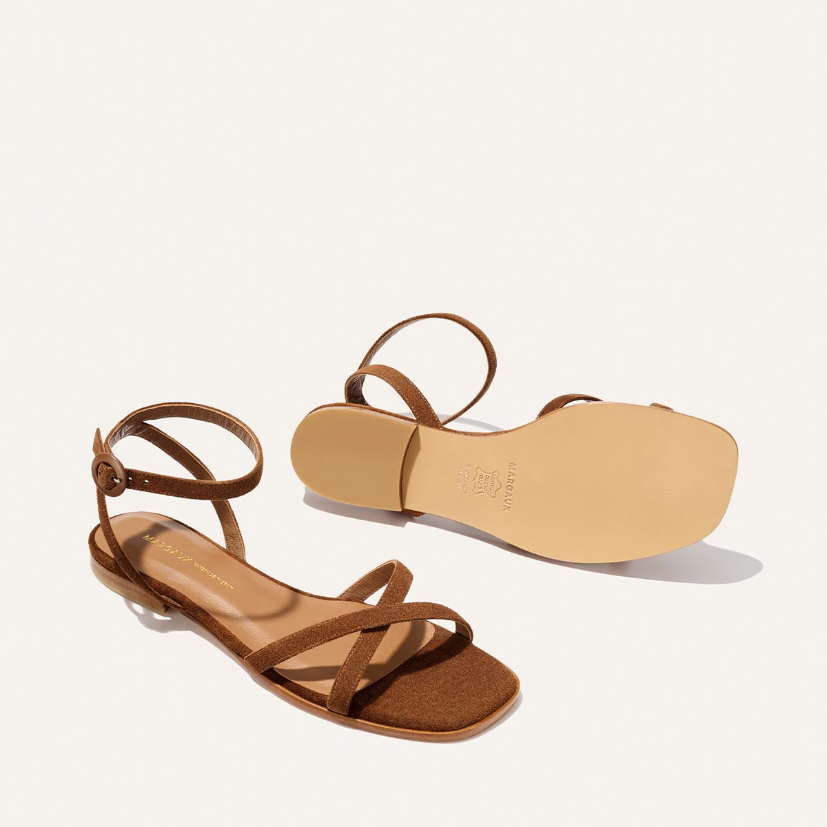 The Flat Sandal in Saddle Suede by Margaux—brown barely-there sandals with ankle straps, low heels, and a foam-cushioned insole—are shown from side and bottom views on a plain white background.
