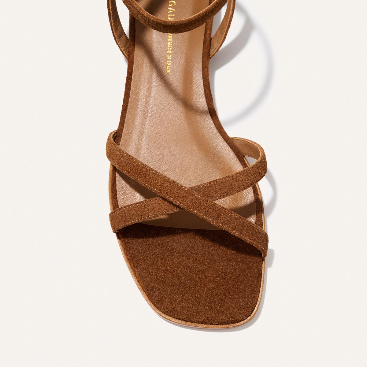 A close-up top view of Margaux's The Flat Sandal in Saddle Suede, featuring thin crisscross straps, a square open toe, and cushioned insole, shown on a light background.