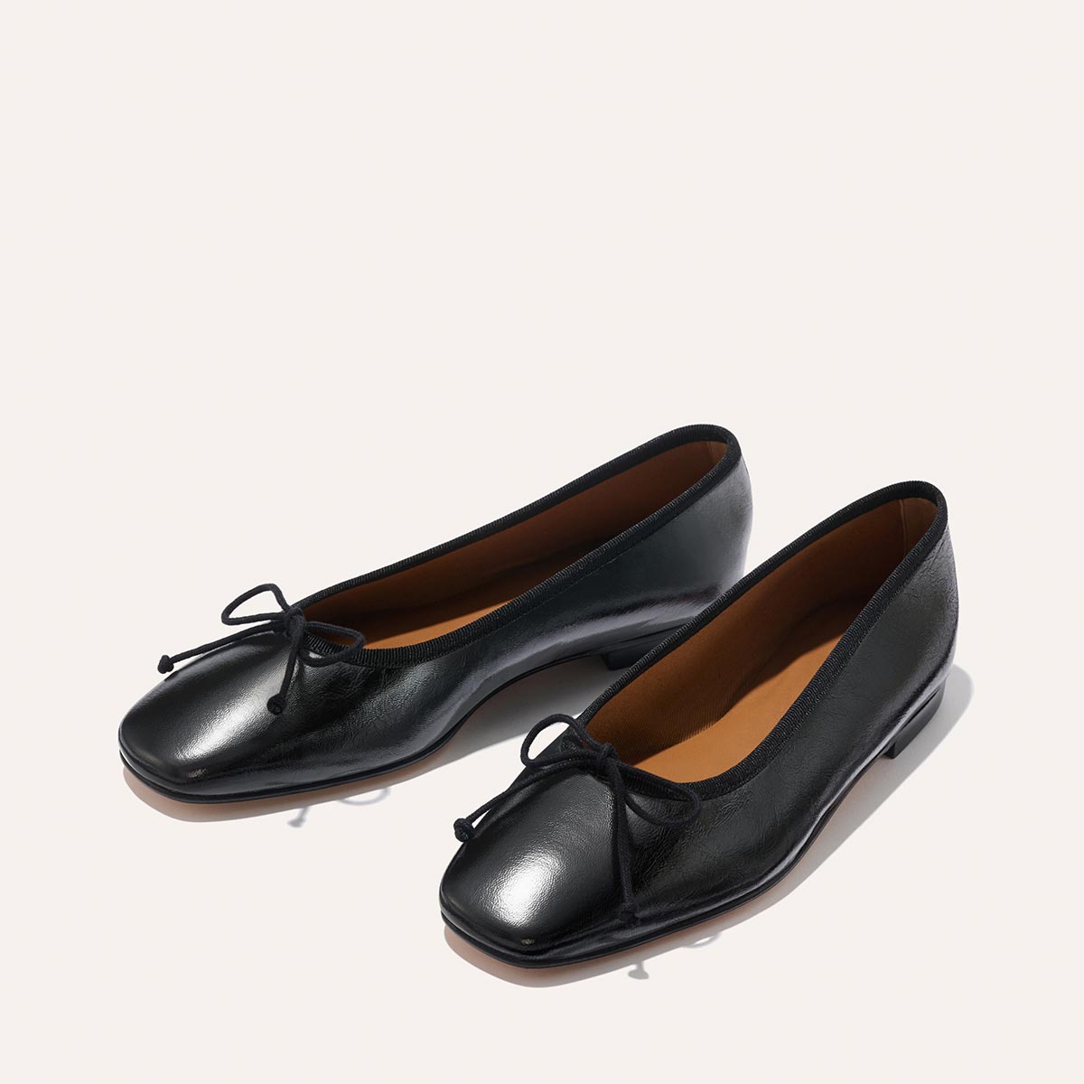 The Fonteyn ballet flats by Margaux feature black crinkle-gloss nappa leather, square toes, low heels, brown insoles, and delicate front bows, all displayed on a light background.