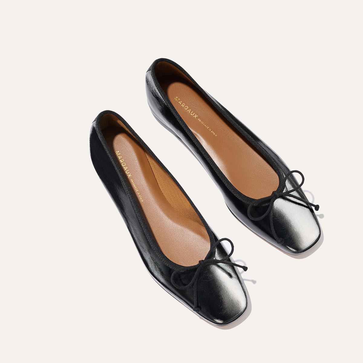 A pair of The Fonteyn ballet flats by Margaux in black crinkle-gloss nappa, featuring square toes and black bows, shown from above on a light background. Their tan insoles display the brand name in gold lettering.