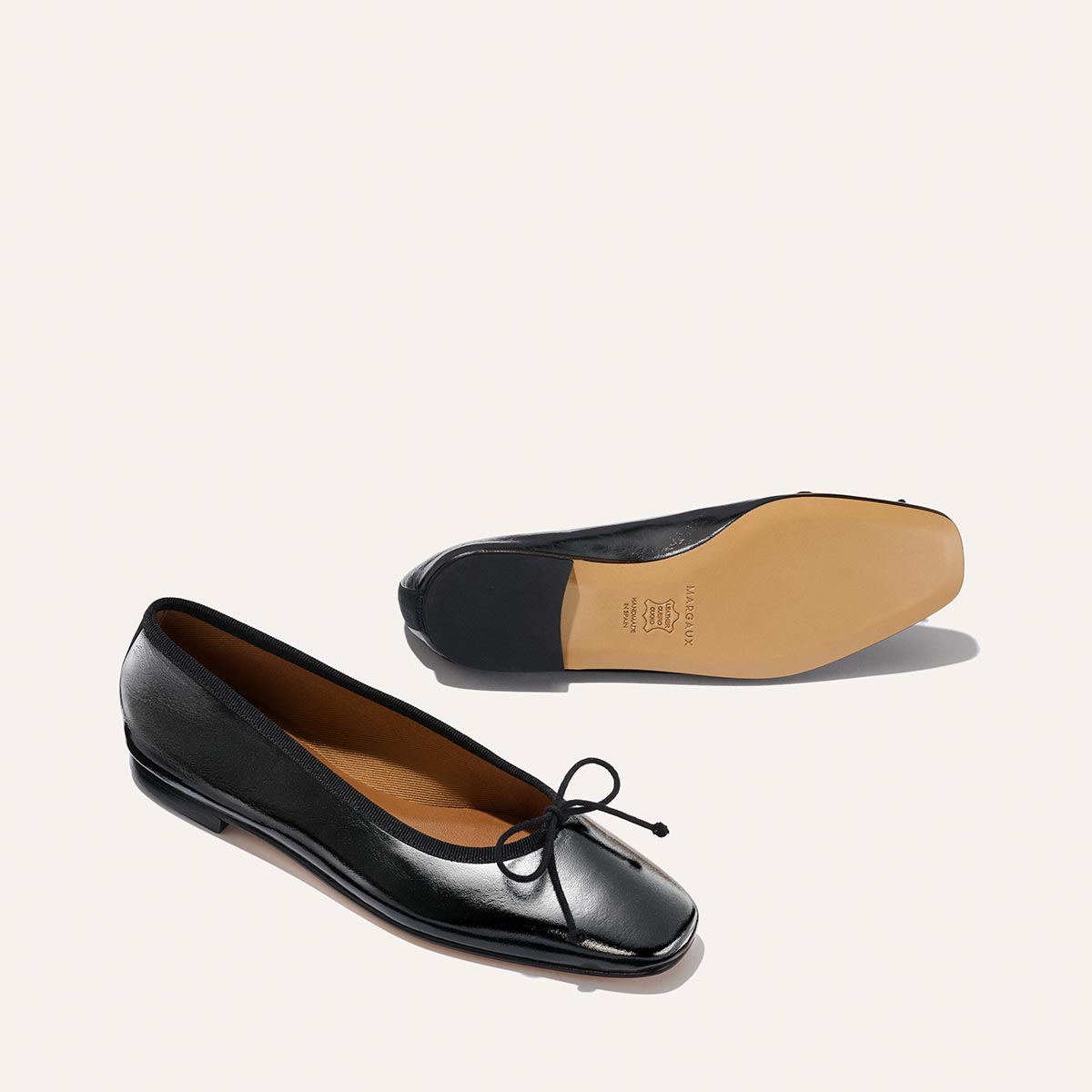 A pair of Margaux The Fonteyn flats in black crinkle-gloss nappa leather, featuring a small bow. One shoe is upright to show the top and side; the other is upside down, highlighting the tan sole and heel.