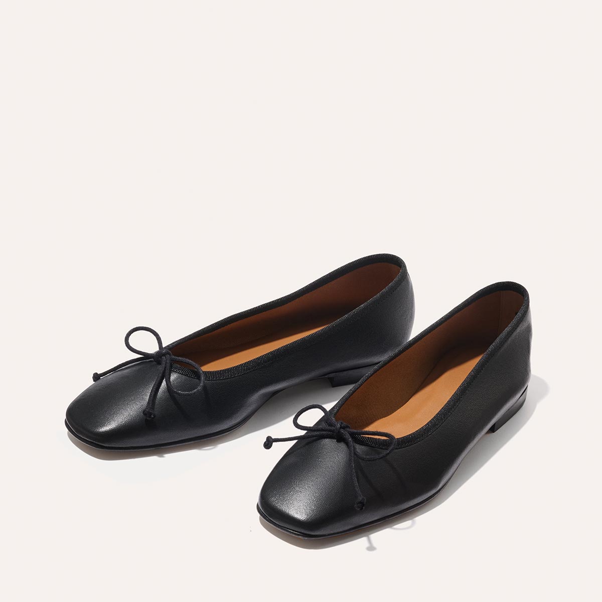 The Fonteyn - Black Nappa ballet flats by Margaux feature handcrafted black leather, square toes, low heels, and small front bows, displayed on a light neutral background.