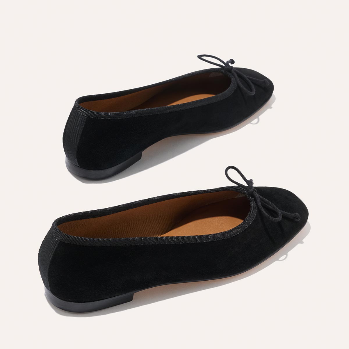 Two pairs of Margaux’s The Fonteyn - Black Suede ballet flats, featuring small front bows, tan insoles, and low black heels, are displayed on a light background with one comfort-first shoe positioned above and slightly behind the other.