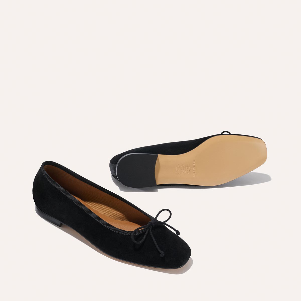 Margaux's The Fonteyn - Black Suede: handcrafted ballet flats with small toe bows. One shoe is upright showing side and top; the other flipped to reveal its light tan sole and heel, both set on an off-white background.