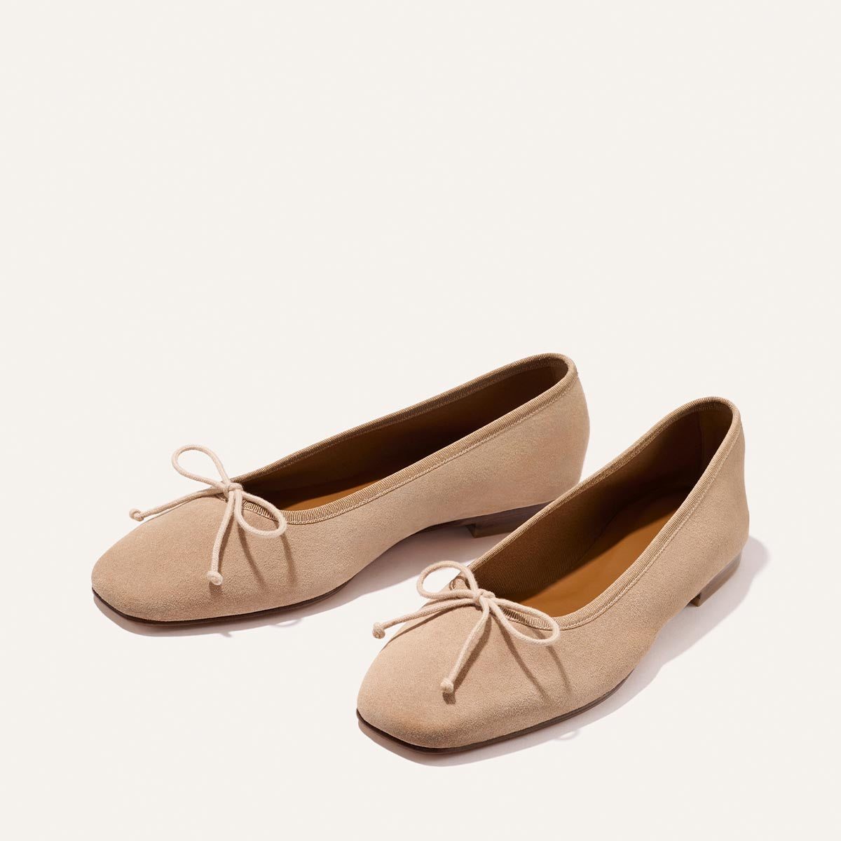 The Fonteyn - Cameo Suede by Margaux: beige suede ballet flats with low heels and decorative bows on rounded toes, shown on a plain light background.
