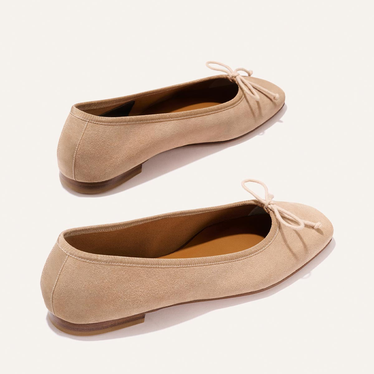 The Fonteyn - Cameo Suede by Margaux: beige suede ballet flats with small bows, rounded toes, and flat soles, displayed on a plain light background.