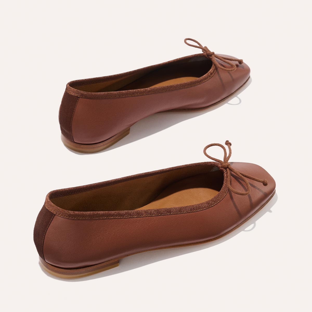 The Fonteyn - Cognac Nappa by Margaux: brown leather ballet flats featuring rounded toes, small bow accents, and brown elastic trim, displayed against a light background.