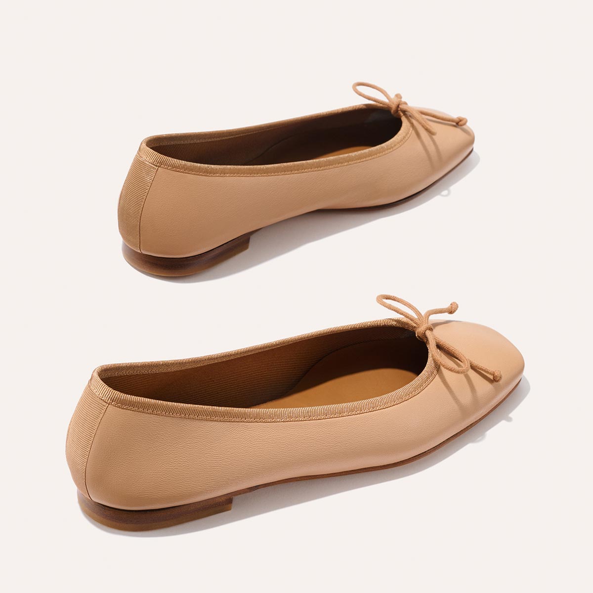 The Margaux Fonteyn - Dune Nappa ballet flats feature tan leather, rounded toes, small front bows, and low wooden heels, displayed against a light background.