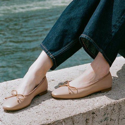 A person in dark blue jeans and Margaux's The Fonteyn ballet flats in Dune Nappa sits on a concrete ledge by the water.