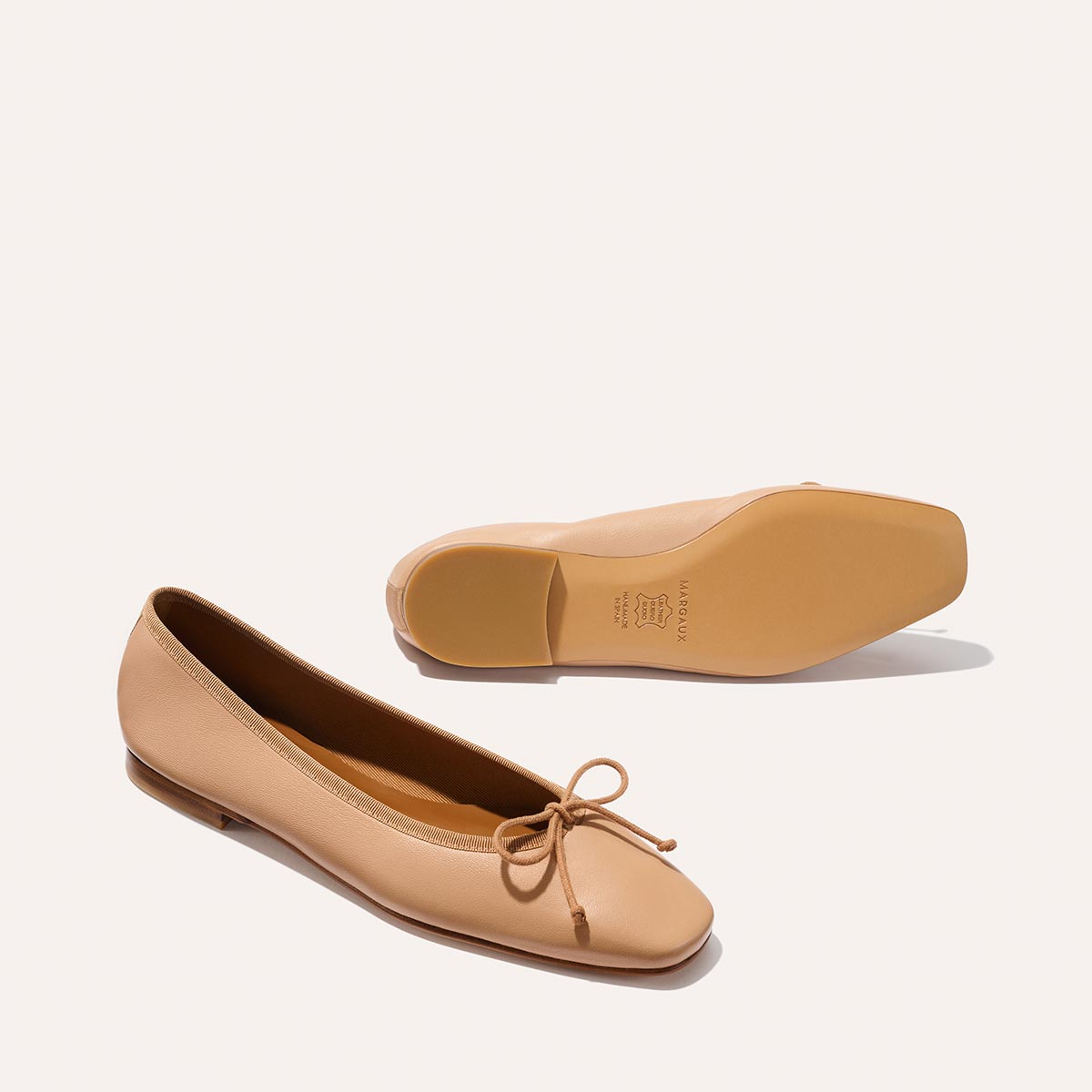 Margaux’s The Fonteyn - Dune Nappa: beige leather ballet flats with a bow detail, shown on a plain light background—one shoe facing up, the other revealing the sole.