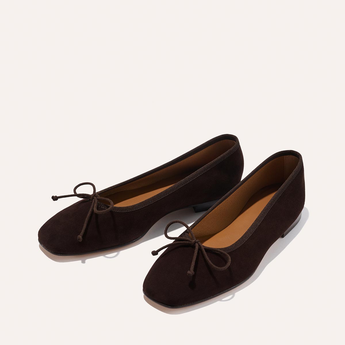 A pair of Margaux's The Fonteyn - Espresso Suede ballet flats, handcrafted in dark brown suede with rounded toes and bow accents, are displayed on a plain light background for timeless comfort.