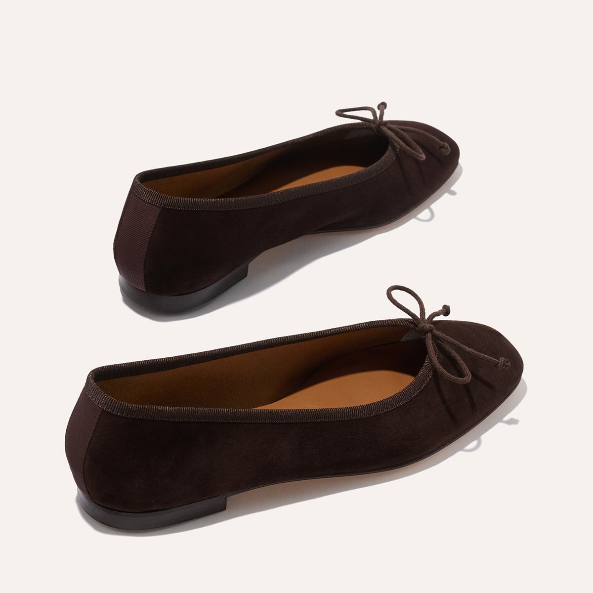 The Fonteyn - Espresso Suede by Margaux are dark brown suede ballet flats with bow details and almond-shaped toes, designed for daily comfort. Handcrafted with flat soles, they're shown from a slightly elevated angle on a light background.