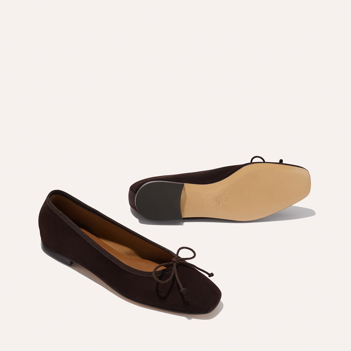 The Fonteyn - Espresso Suede by Margaux: dark brown suede ballet flats with petite front bows, designed for comfort. One shoe stands upright; the other displays its beige sole and low heel against a simple light backdrop.
