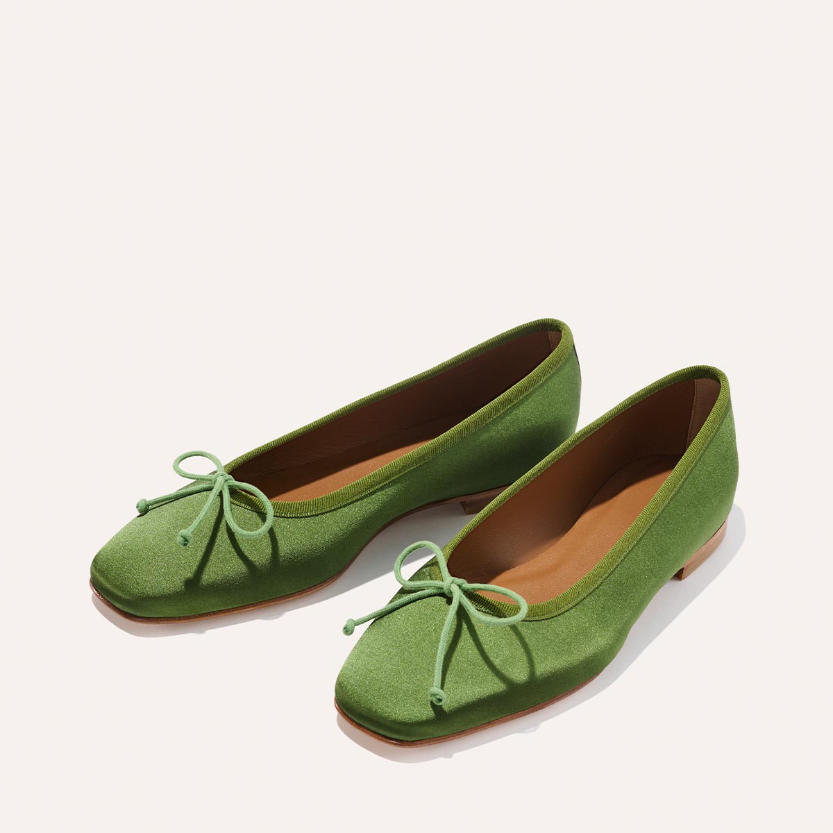 The Fonteyn - Forest Satin by Margaux: green satin ballet flats with square toes, delicate front bows, brown soles, and comfort-first craftsmanship, displayed on a light background.