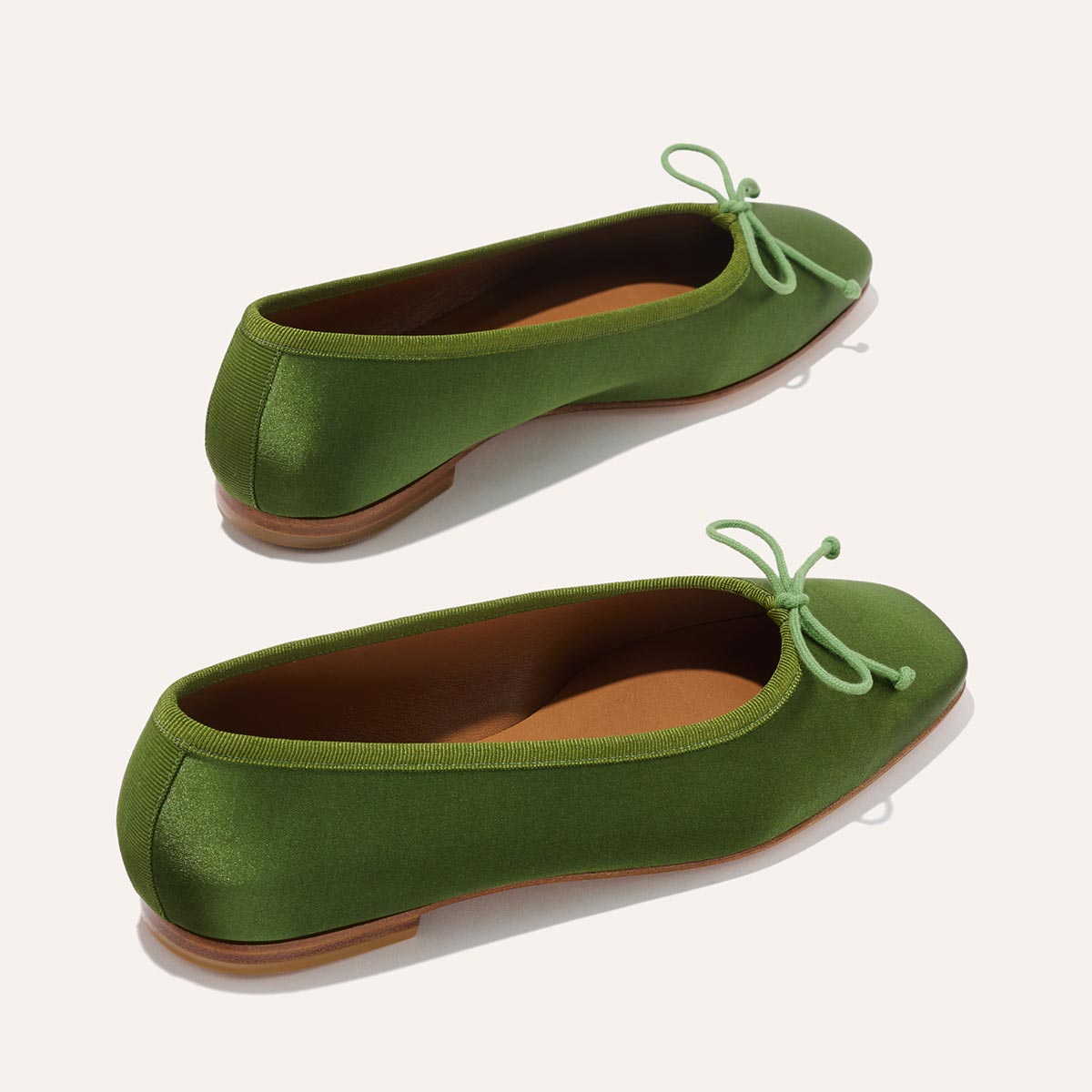 The Fonteyn - Forest Satin by Margaux: green ballet flats with rounded toes, matching bows, tan inner soles, and brown outer soles—crafted for comfort and stylishly displayed against a light backdrop.