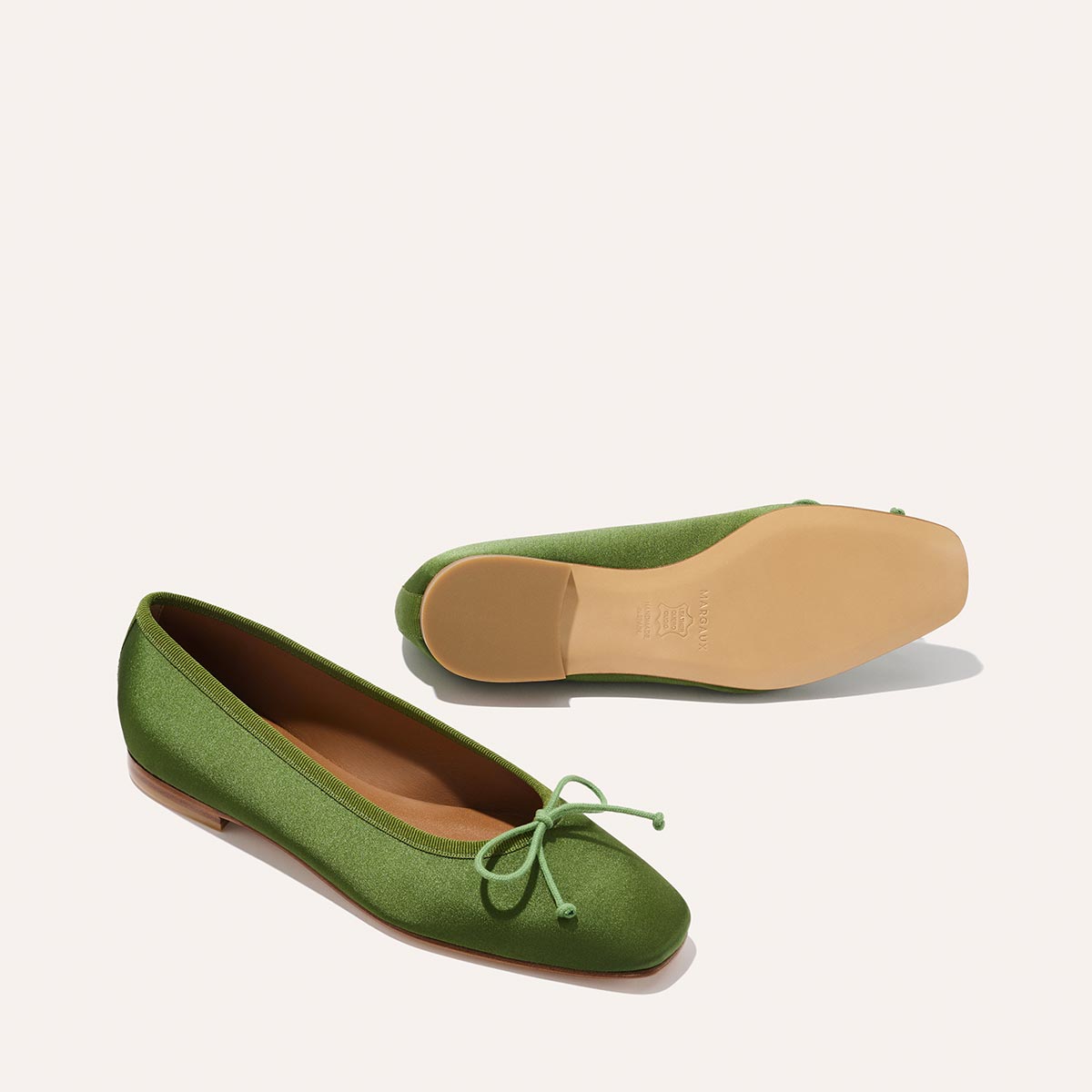 The Fonteyn - Forest Satin by Margaux: a pair of handcrafted green ballet flats with small front bows, shown one upright and one on its side to display the tan sole, set against a plain light background.