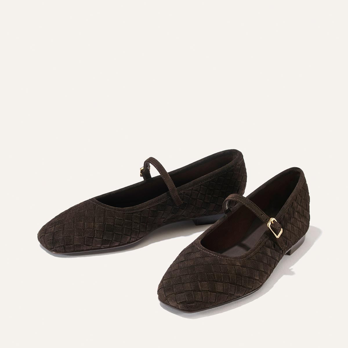 A pair of Margaux’s The Fonteyn Jane flats in dark chocolate woven Pasado suede, featuring gold buckles on the Mary Jane straps, displayed against a plain, light background.