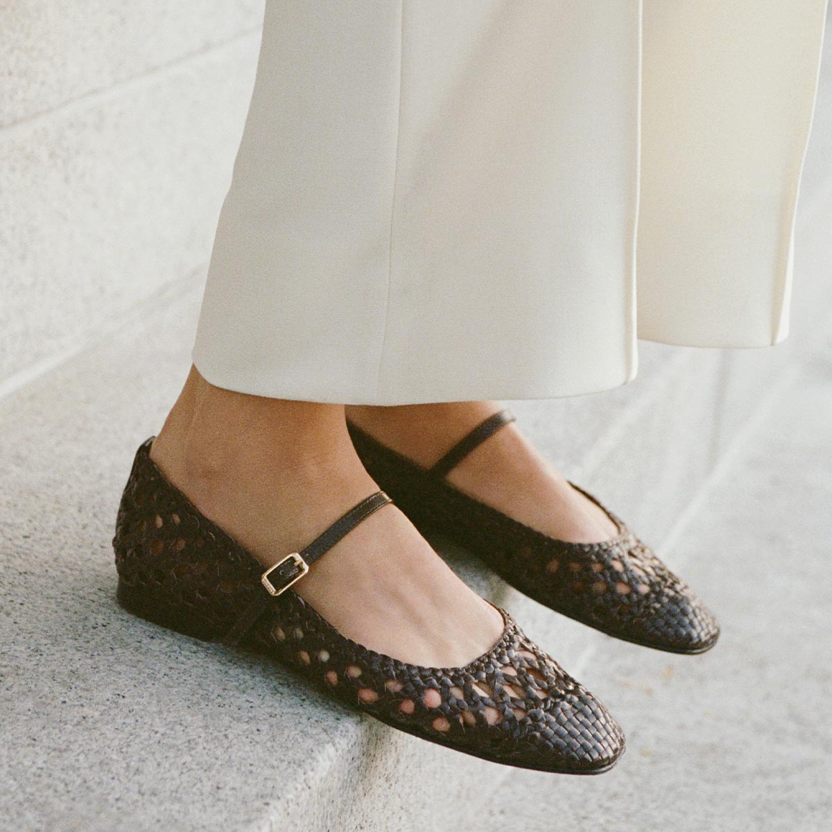 A person wearing cream-colored pants and Margaux's The Fonteyn Jane - Espresso Woven Leather flats, featuring an adjustable strap and small buckle, stands on a concrete surface.