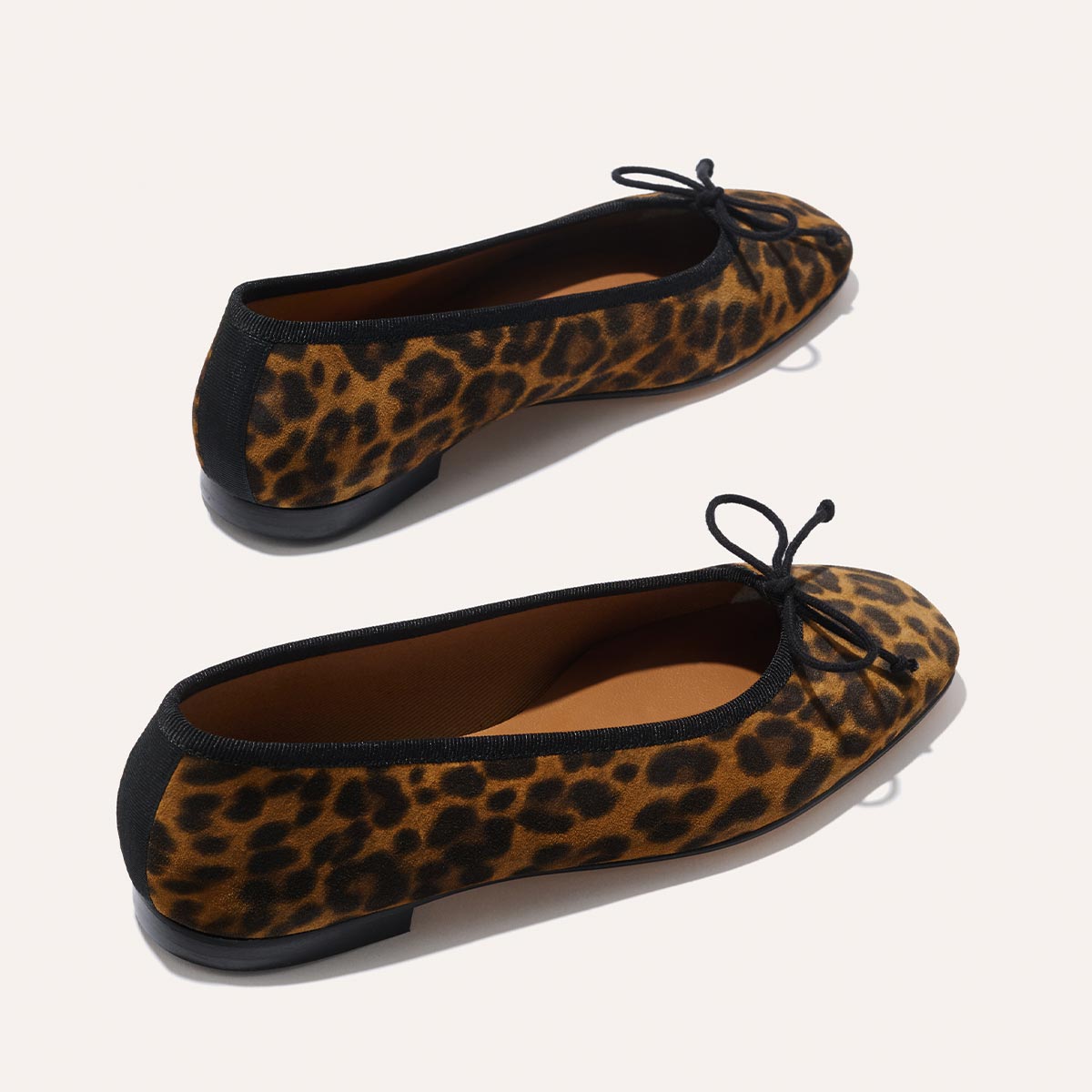 Margauxs The Fonteyn - Leopard Suede ballet flats feature a leopard print, black trim, black bow at the toe, and flat black soles, shown on a light background.