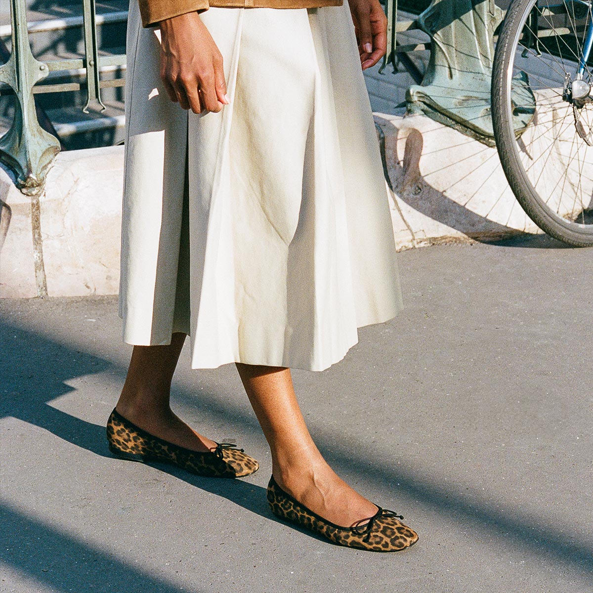 A person wearing a cream-colored midi skirt and Margaux’s The Fonteyn ballet flats in Leopard Suede stands on a sunlit sidewalk near a bicycle.