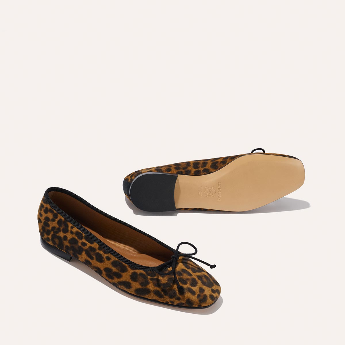 The Fonteyn - Leopard Suede ballet flats by Margaux are displayed against a plain background: one shows the leopard print and black bow on top, while the other reveals the tan sole and black heel.