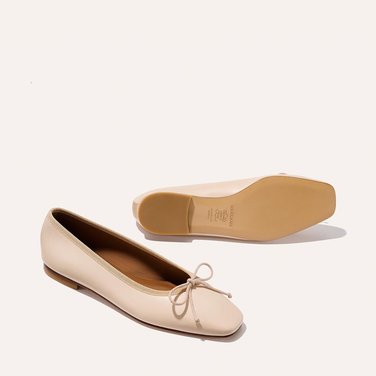 A pair of Margaux’s The Fonteyn - Magnolia Nappa beige ballet flats with a small bow; one shoe is upright showing the top, and the other is flipped to display the sole against a plain background.