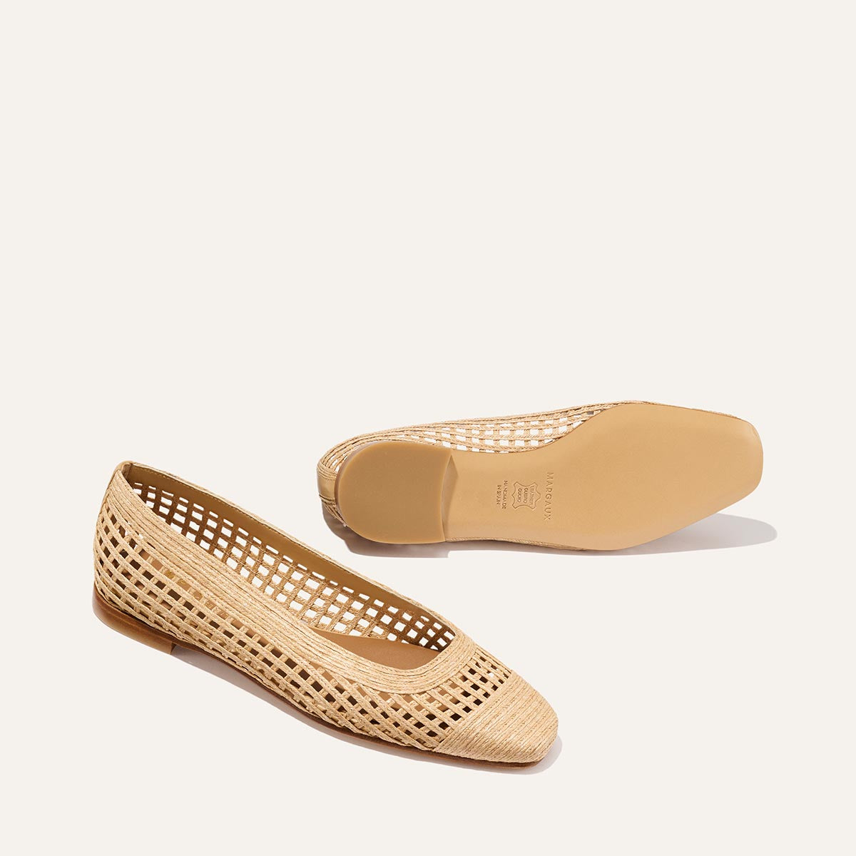 The Fonteyn - Natural Grid Raffia by Margaux features beige woven flats with a lattice design; one shoe stands upright to show the side and top, while the other is flipped to reveal the smooth sole—ideal for elevating any look.