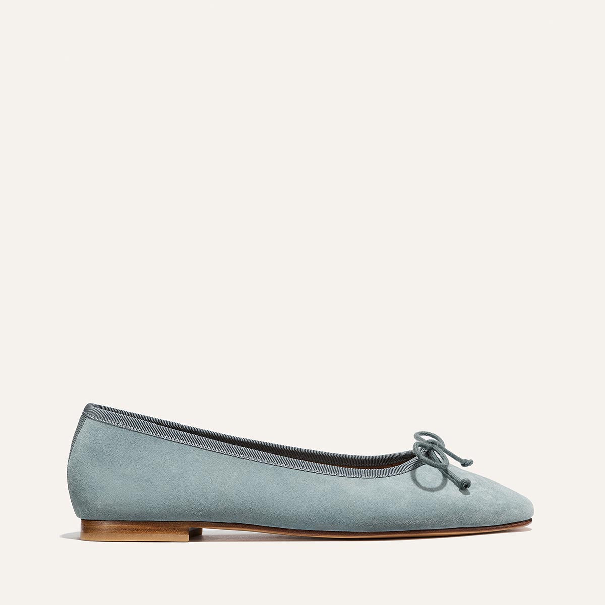 Margaux’s The Fonteyn - Ocean Suede is a comfort-focused light blue suede ballet flat with a small front bow, dark grey trim, and a low wooden heel, showcased on an off-white background.