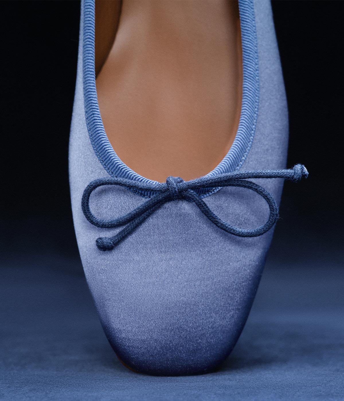 A close-up of the front of a blue satin ballet flat shoe with a cord bow on top, against a dark background.