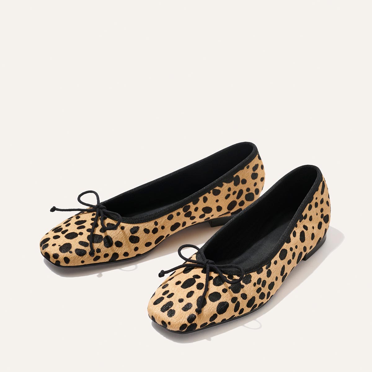 Experience timeless comfort and style with Margaux’s The Fonteyn in Tan Cheetah Haircalf—a pair of elegant ballet flats featuring a soft beige base, classic black accents, and playful black bows on the toes, perfect for effortless everyday wear.