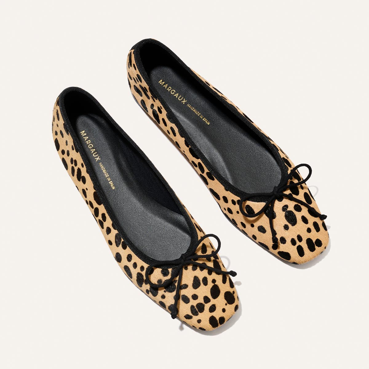 A stylish pair of The Fonteyn - Tan Cheetah Haircalf ballet flats from Margaux, featuring a chic animal print with black trim and bows, displayed against a light background. Crafted for comfort, the insoles are stamped with the Margaux brand name.