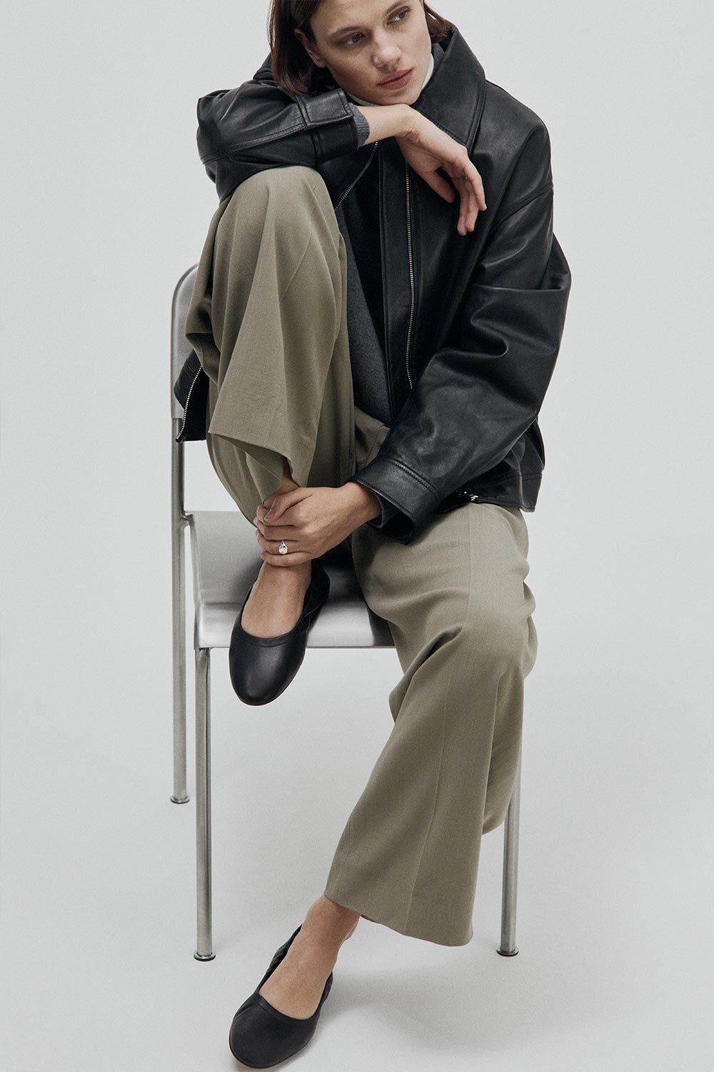 Woman sitting on a chair wearing a black leather jacket, beige pants, and black shoes.