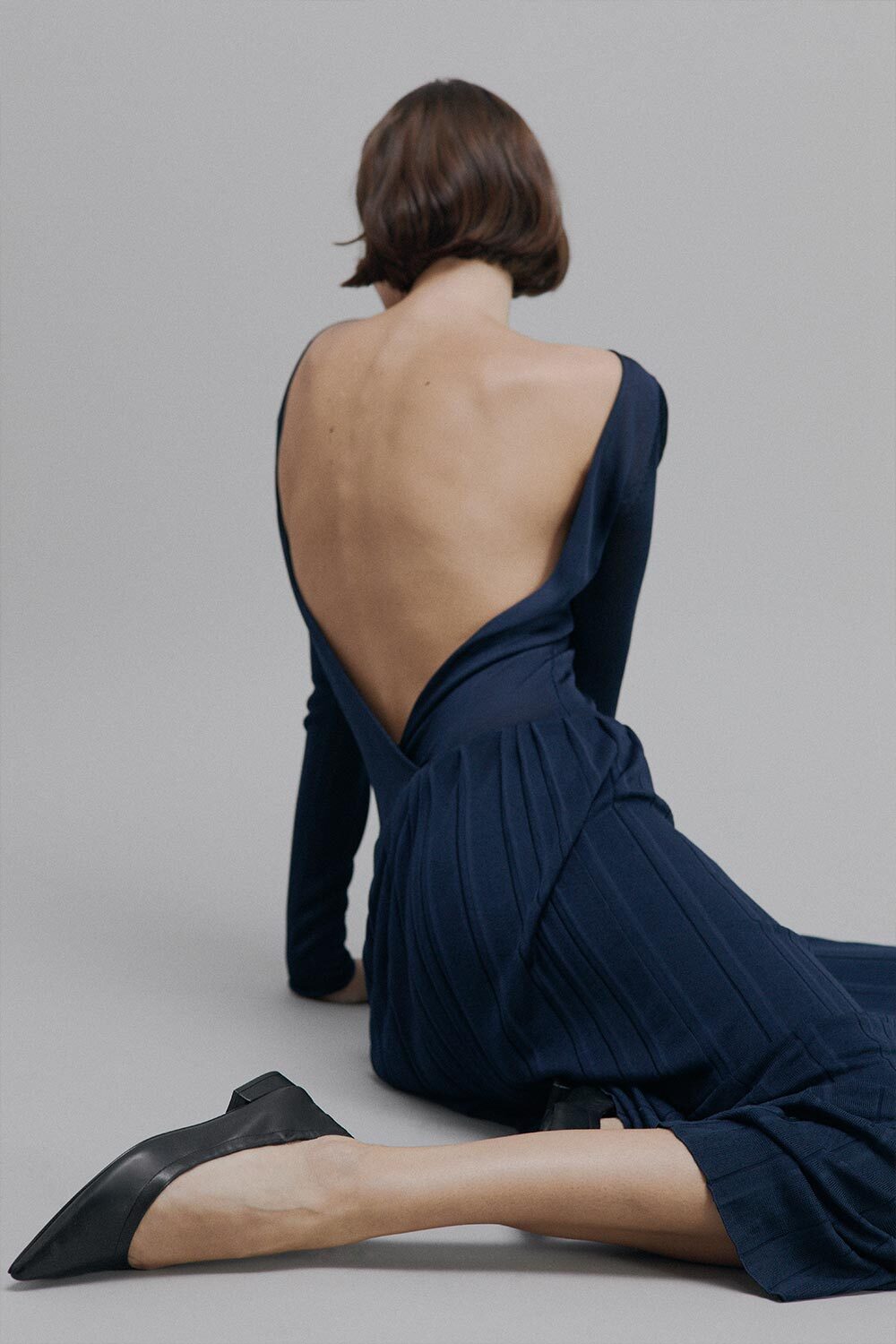 Woman wearing a navy blue dress with a backless design, sitting on a gray floor.