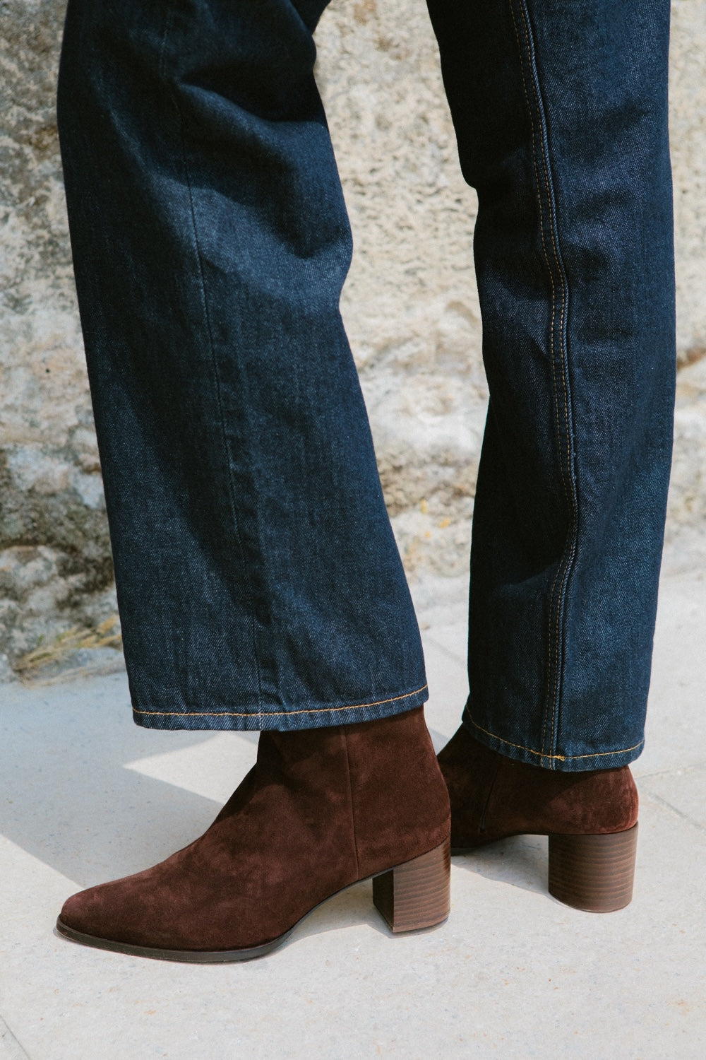 Brown suede boots worn with blue jeans against a stone wall background
