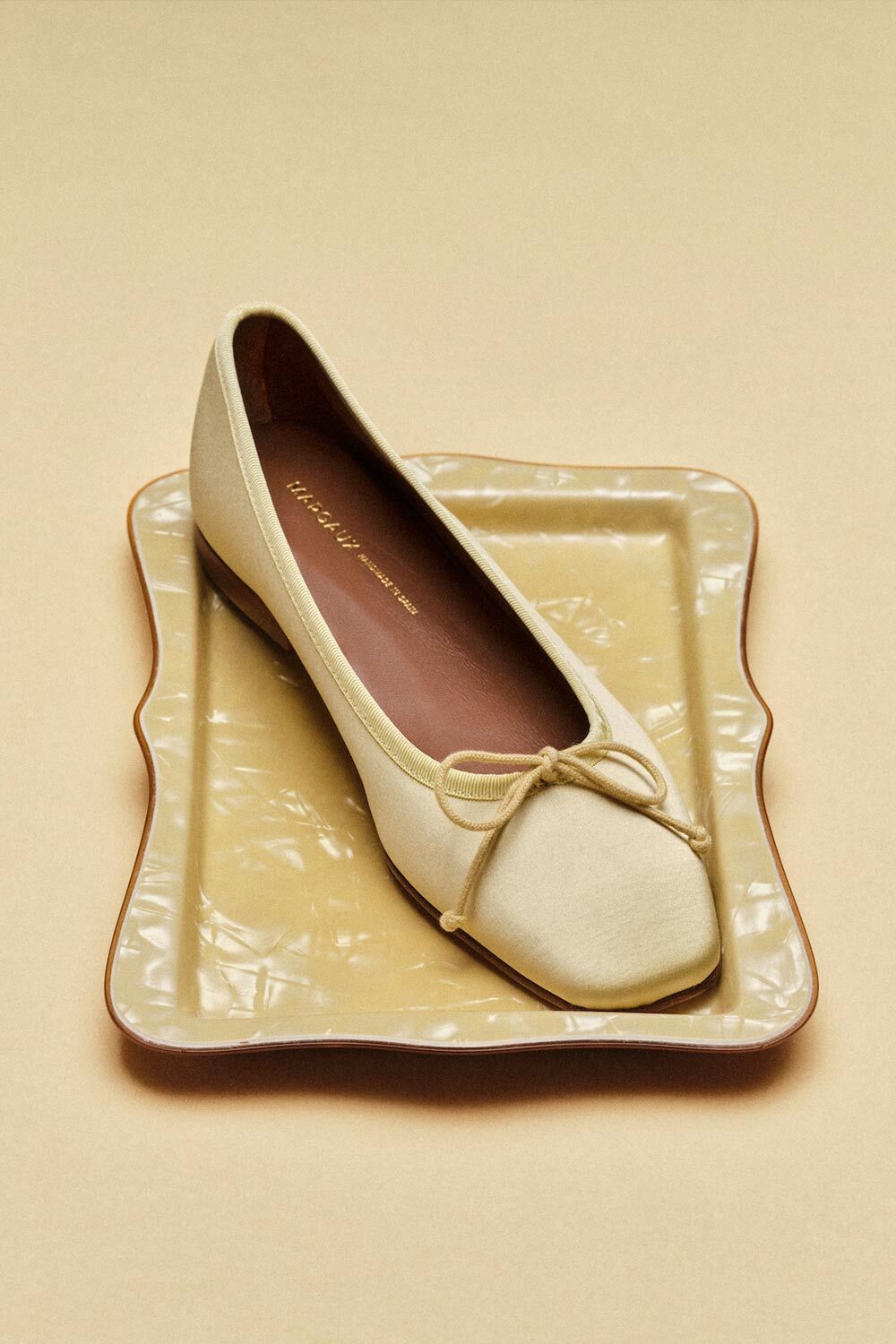 A cream-colored ballet flat with a small bow on the toe rests on a beige, wavy-edged ceramic tray against a matching beige background.