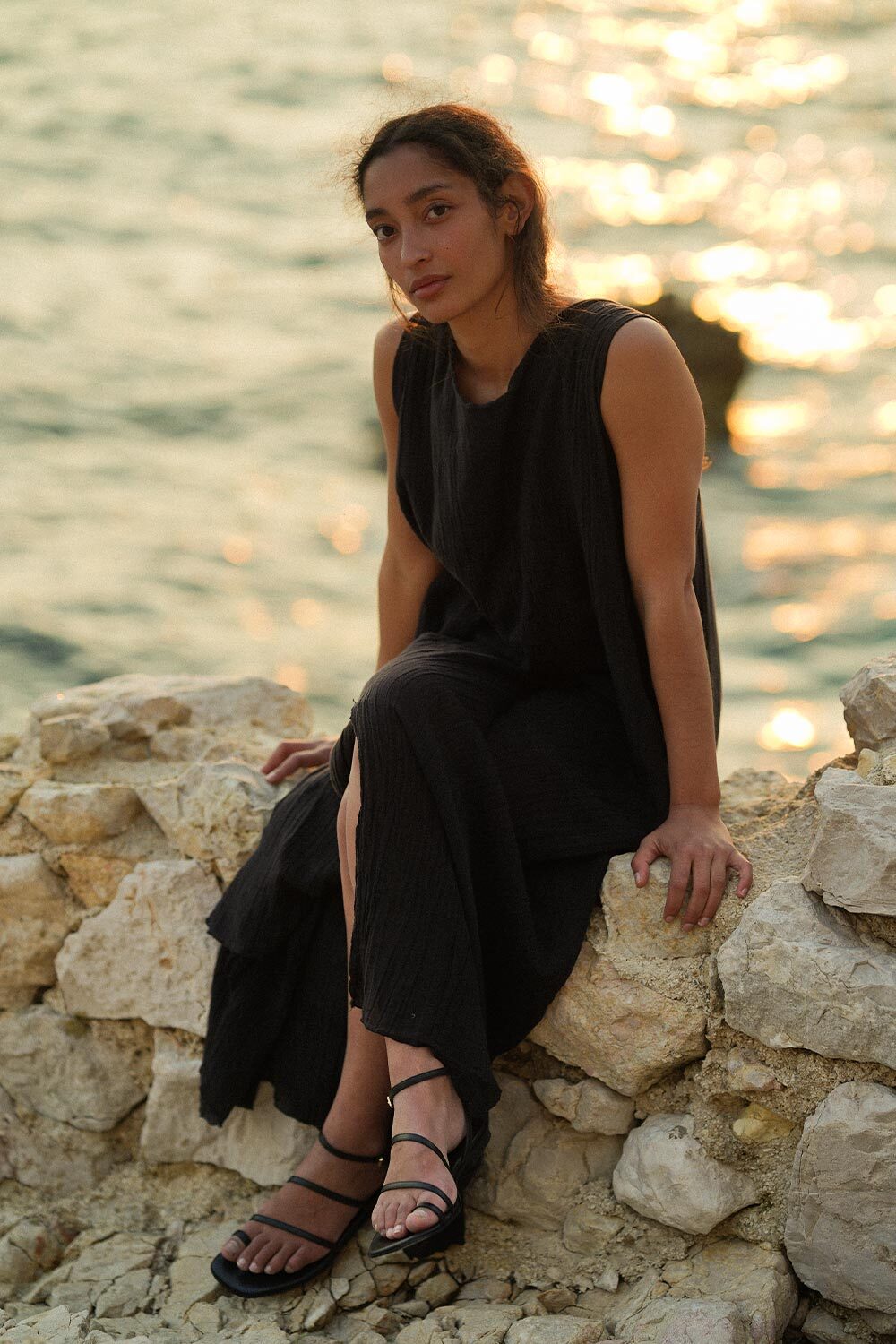 A woman in a sleeveless black dress sits on light-colored rocks by the water at sunset, with soft sunlight reflecting on the waves behind her.