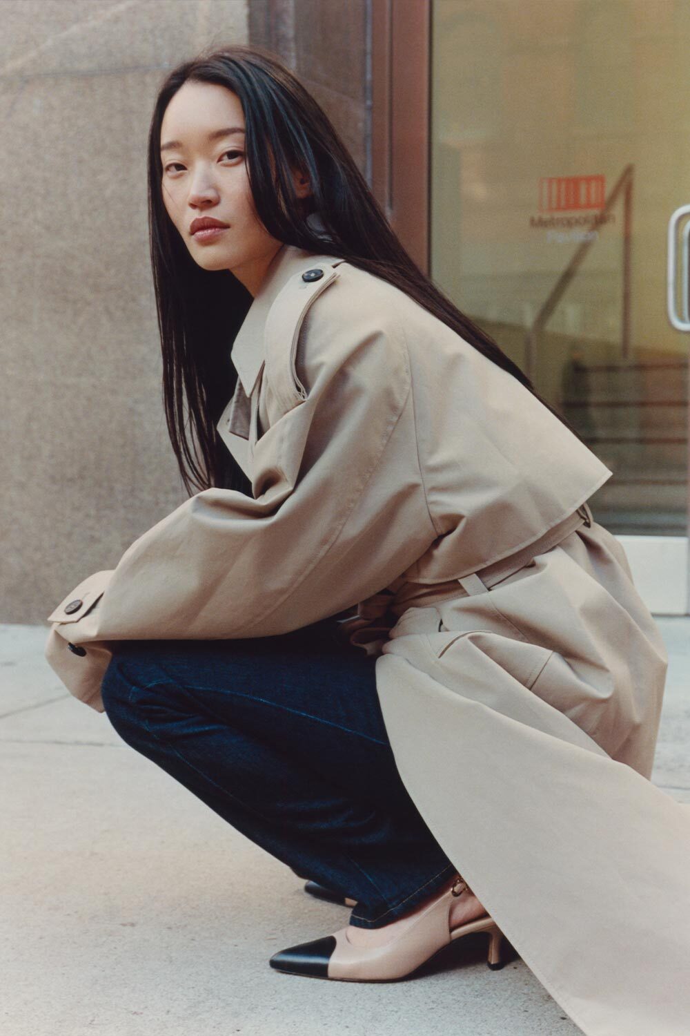 Woman in a beige trench coat squatting on a sidewalk