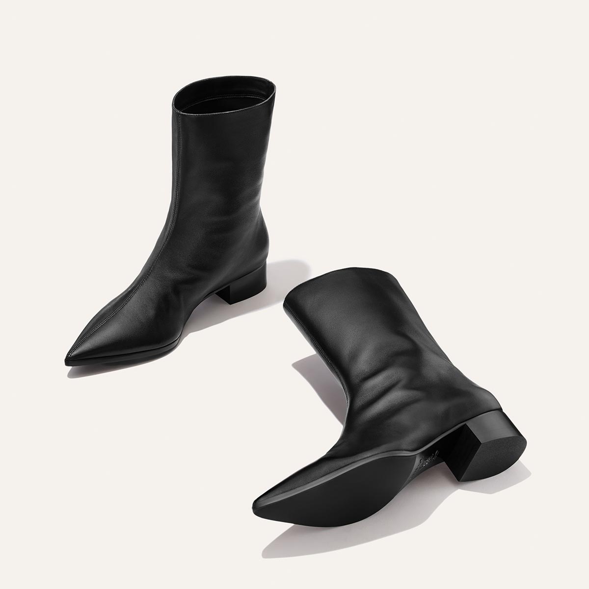 The Hayes Boot in Black Nappa by Margaux, featuring a mid-calf height, pointed toe, and low block heel in smooth leather, is displayed against a light background. One boot stands upright while the other rests on its side.